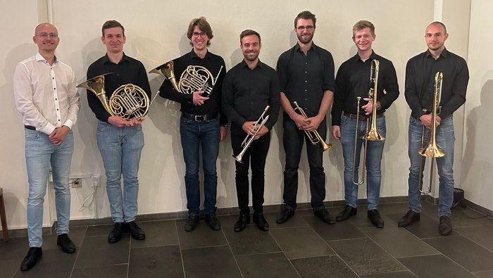 Sieben lächelnde Männer halten Blechblasinstrumente im Innenraum.
